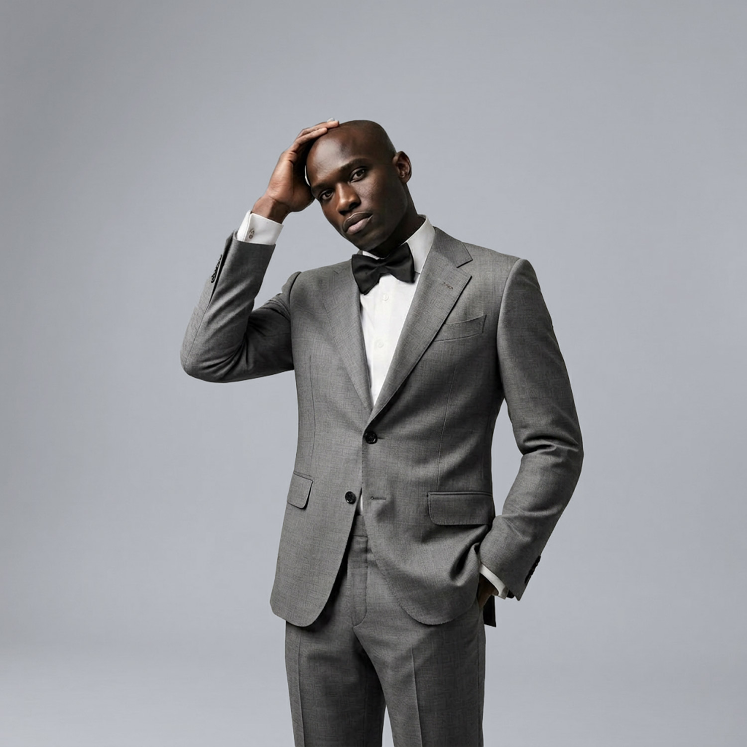 Man in tailored grey suit with bow tie posing with hand on head against a clean studio background