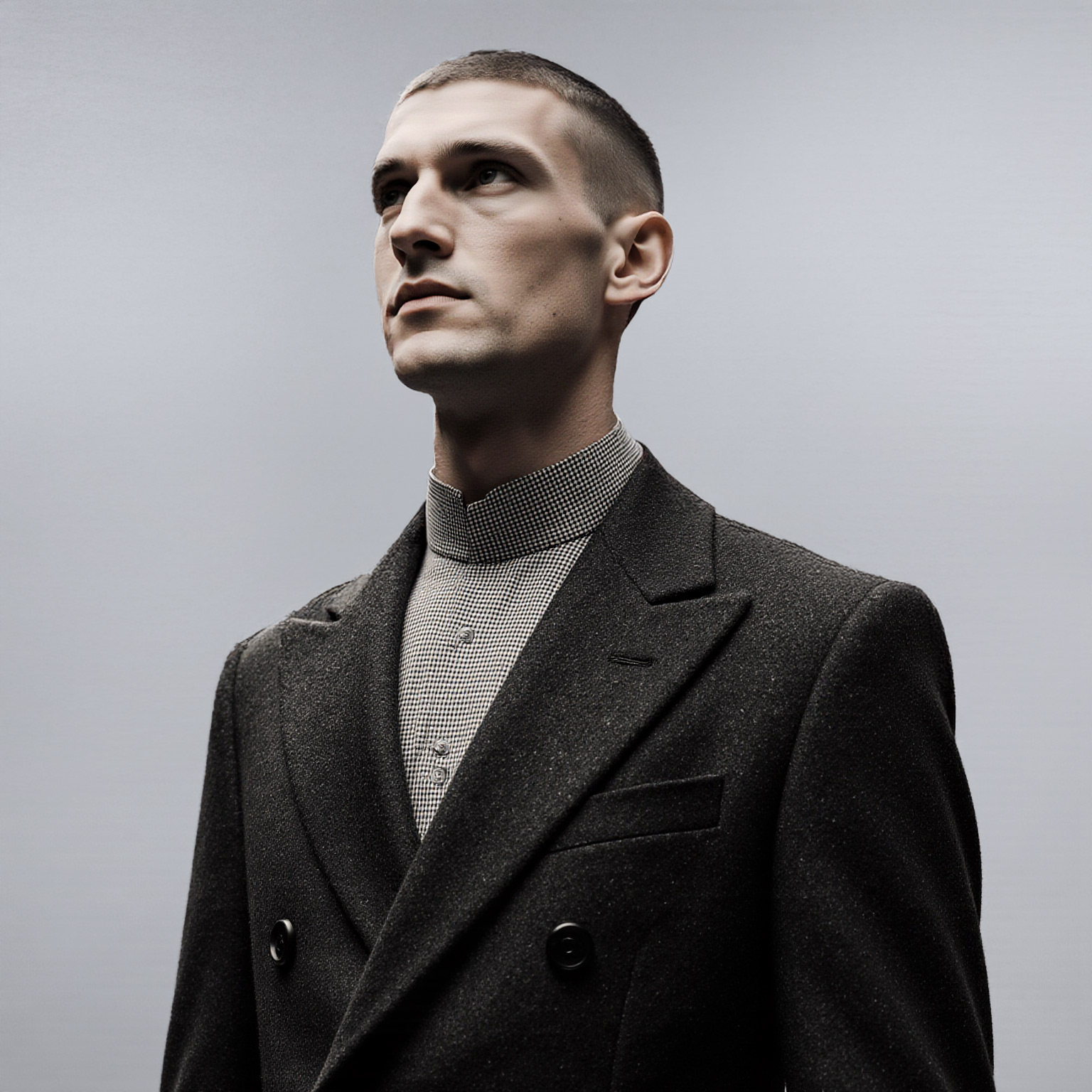 Man in structured double-breasted coat with band collar shirt looking upward in a studio setting