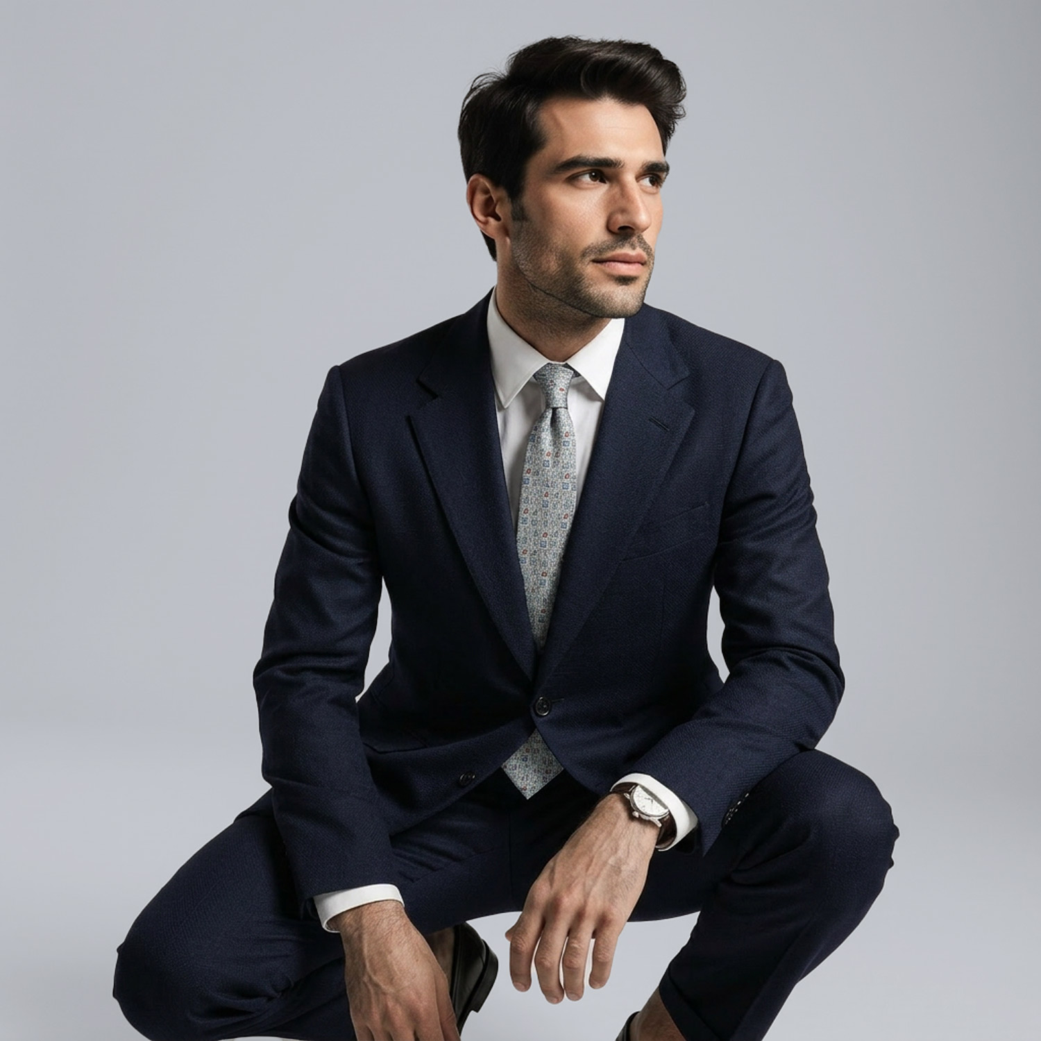 Man in navy tailored suit crouching with tie and watch against a clean studio background