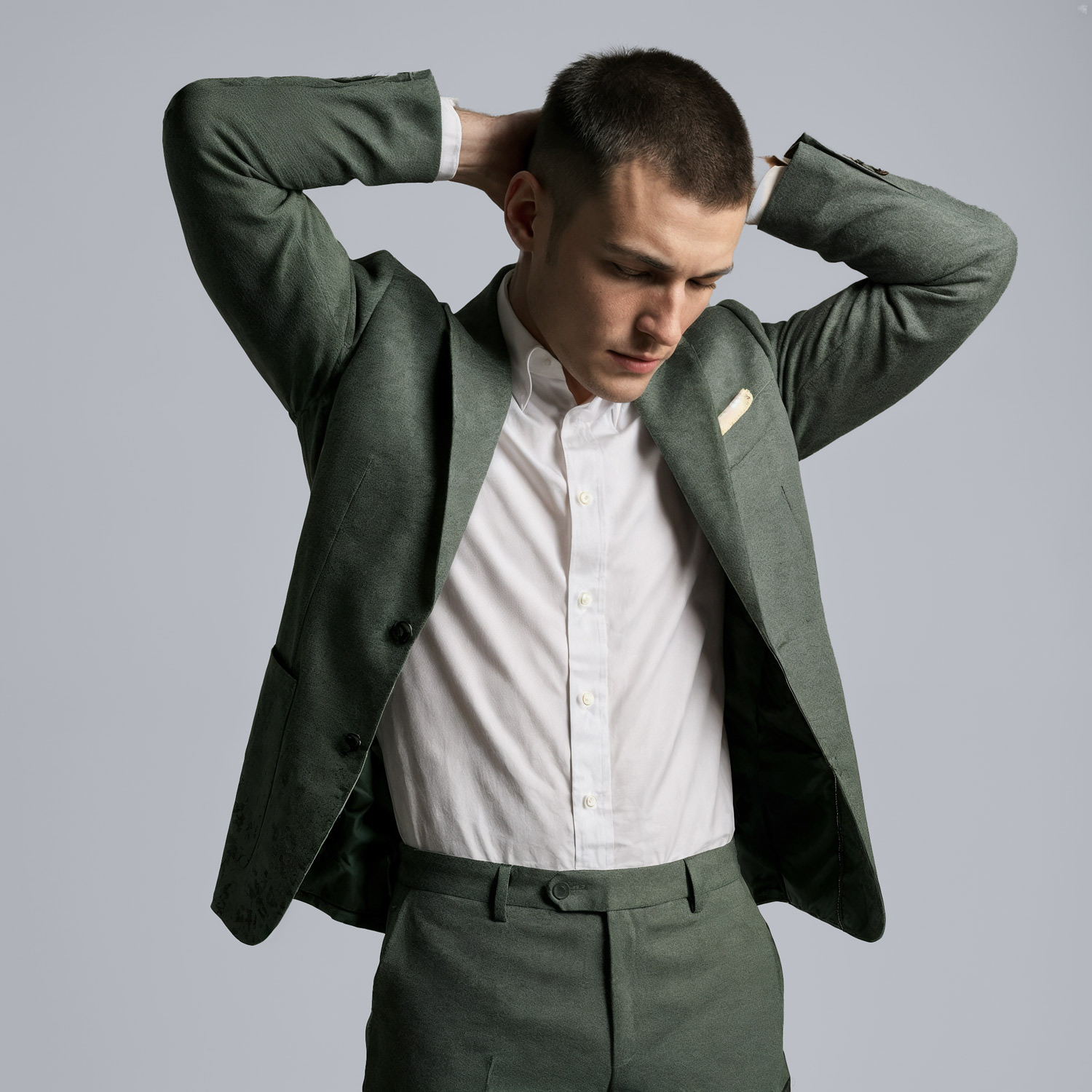 Man in green tailored suit adjusting jacket with hands behind head in a studio setting