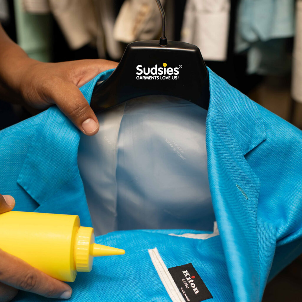 Sudsies garment care professional treating blue blazer with cleaning solution on hanger